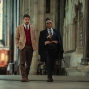 "Two well-dressed men walking together inside a grand, historic stone building with high ceilings and stained glass windows, suggesting a church or cathedral setting."