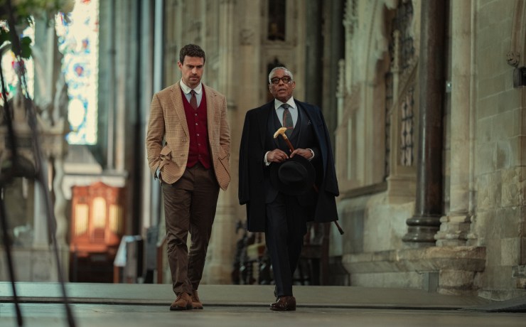 "Two well-dressed men walking together inside a grand, historic stone building with high ceilings and stained glass windows, suggesting a church or cathedral setting."