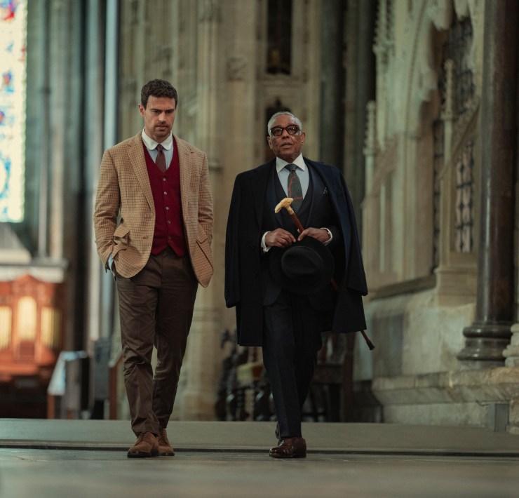 "Two well-dressed men walking together inside a grand, historic stone building with high ceilings and stained glass windows, suggesting a church or cathedral setting."