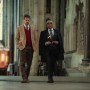 "Two well-dressed men walking together inside a grand, historic stone building with high ceilings and stained glass windows, suggesting a church or cathedral setting."